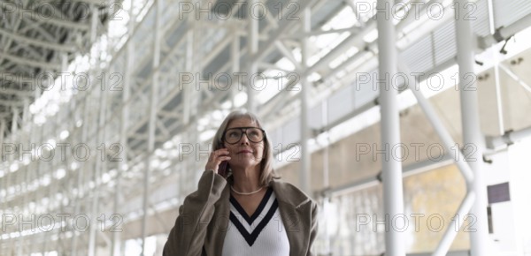A middle-aged businesswoman with glasses is engaged in a phone conversation while strolling through a bright and spacious airport terminal. Her confident demeanor suggests professionalism and purpose