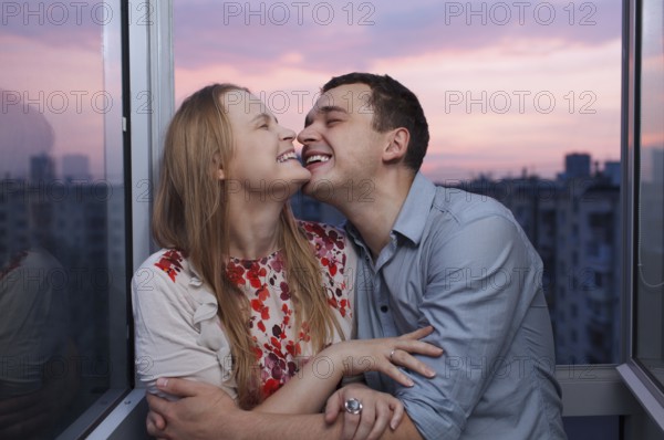 Playful young couple embracing and laughing on the balcony at a high floor during sunset