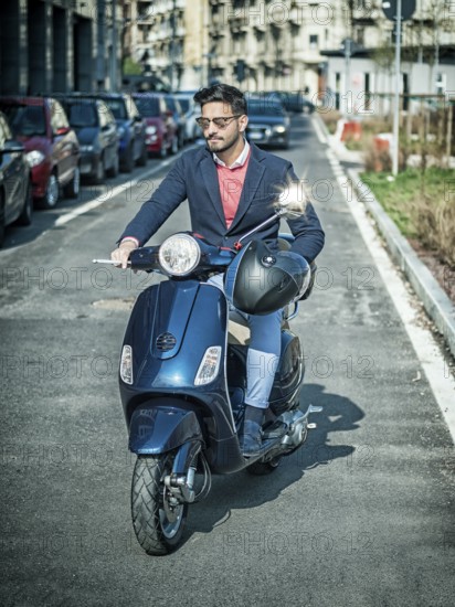 Young stylish man in suit and sunglasses driving scooter on empty road in sunny city