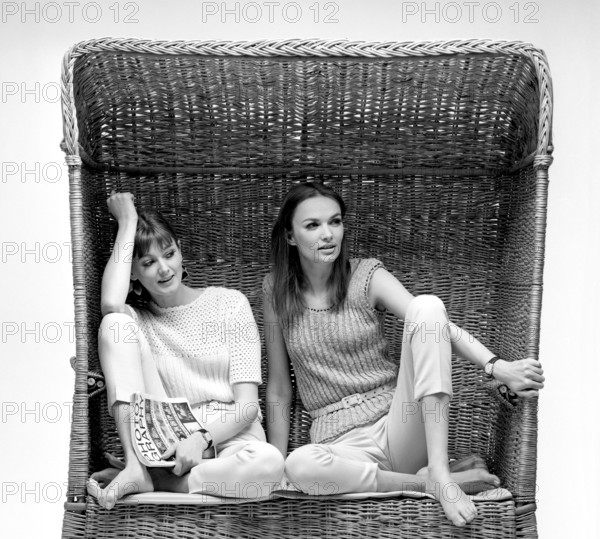 In the 1960s. Two fashion models, in typical 1960s summer outfits photographed in a studio wearing knitted sweaters and trousers. They are sitting in a wicker chair usually a type of furniture a accosiated with a beach or a terrace. 1966