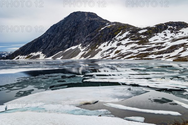 The very cold clear water of the lake Djupvatnet is covered with ice in July. The mountains are covered with freshly fallen snow. Kingdom of the Snow Queen. Magical places in Norway
