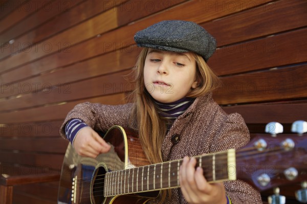 Blond kid girl playing guitar with winter beret and coat on wooden background