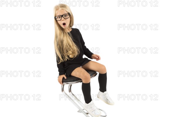 Isolated on white, pretty caucasian blonde little girl in black dress, glasses, black socks and white shoes sitting, look at camera, amazed