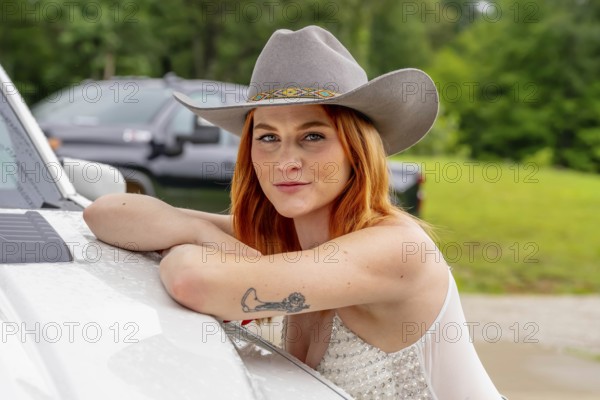 A beautiful redhead model poses in a country setting wearing cowgirl attire against her truck