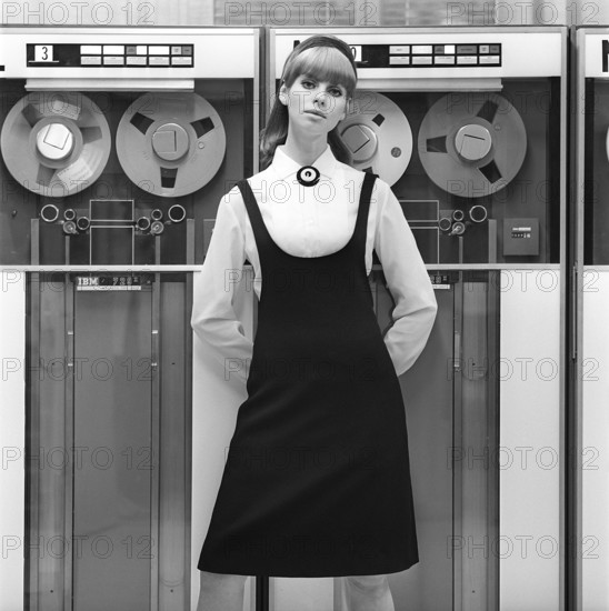 In the 1960s. Fashion model wearing the typical clothes of 1965. The photograph is taken in a 1960s mainframe computer room with magnetic tape units in use where the tape containing information is being read or entered. She is wearing a white shirt and a black dress over it. Picture taken 1965. ref DV16