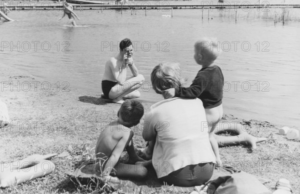 Family in the 1950s. A mother and her children sits on the beach while her husband is filming her with a amateur film camera. The film was then developed and shown on a film projector on a fold up screen. The film had no sound. The film cameras were not battery operated and you had to wind them up like a clock to make them work. Sweden 1953
