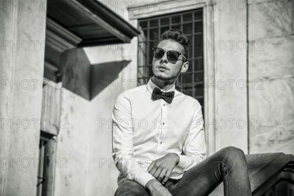 Attractive young man sitting next to walls in city, looking away, wearing sunglasses. Half body shot
