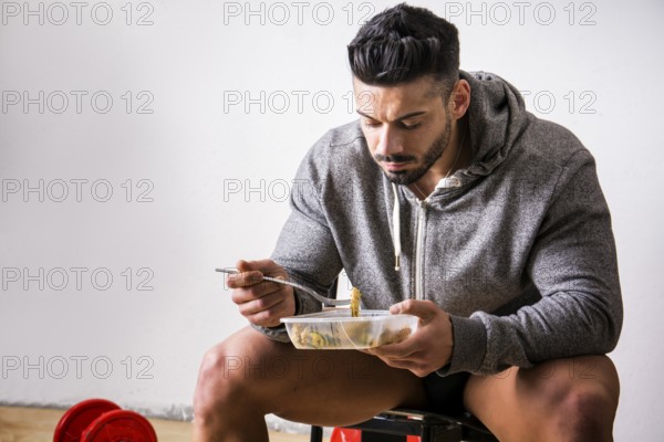 Hungry muscular young man gulping down food looking at plastic container without pausing as he takes another mouthful