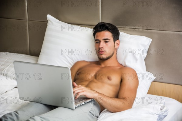 Attractive Young Shirtless Muscular Man with Serious Expression, Reclining Comfortably with Laptop on Bed Working on his Start-up Business - Young Male College or University Student Doing Homework, in Bedroom
