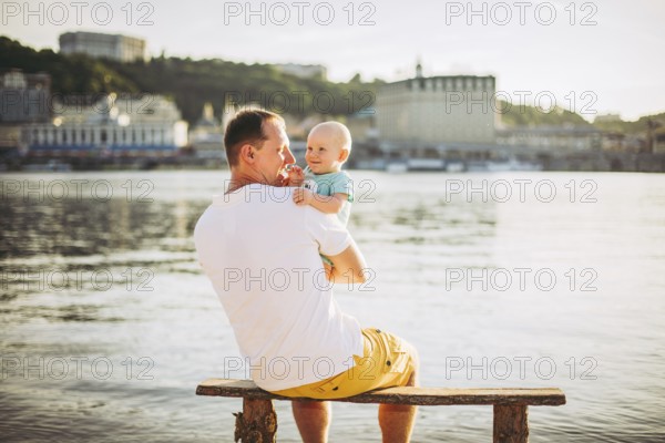 Subject recreation father and little son. Young caucasian dad sits on wooden bench overlooking the town Kiev and holding river of the Dnieper, hugging a toddler baby on sandy beach on the banks pond