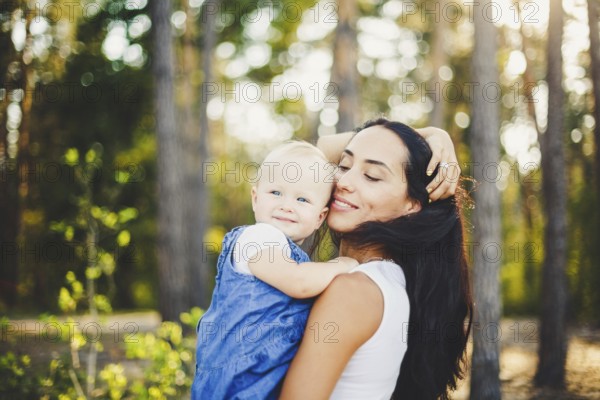 Young beautiful fashionable mother with long brunette hair holding a daughter's blonde with blue eyes one year of birth in a coniferous forest in a summer park. Wearing jeans clothes