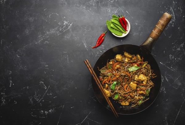 Stir fry soba noodles with chicken, vegetables in old rustic wok pan, chopsticks on black stone background, close up, top view. Traditional asian/thai meal, space for text