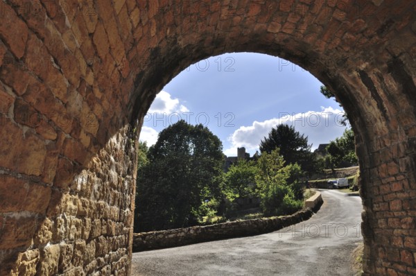 Domme in Perigord, View through a stone archway overlooking a curving path lined with trees under a bright blue sky