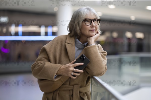 A woman with stylish glasses leans against a railing, thoughtfully holding her smartphone. She appears lost in contemplation in a contemporary setting filled with soft lighting