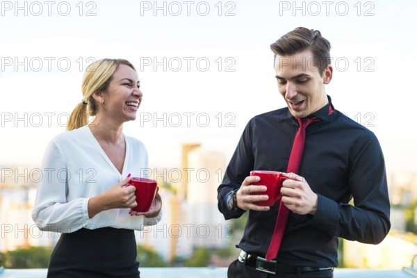 Couple of caucasian businessmen, young attractive brunette man in dark suit, red tie and black shirt, and pretty blonde woman in white blouse and black skirt stand on the roof with red cups of coffe, having a conversation, guy is shy blured city on background