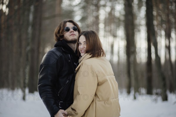 The couple stands happily in a snowy forest, wearing warm clothing and sunglasses