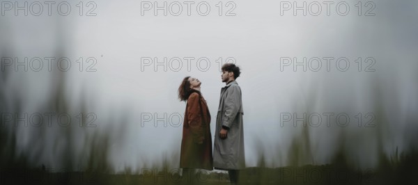 A couple standing in a field, each looking upwards, dressed in long coats with a sense of connection