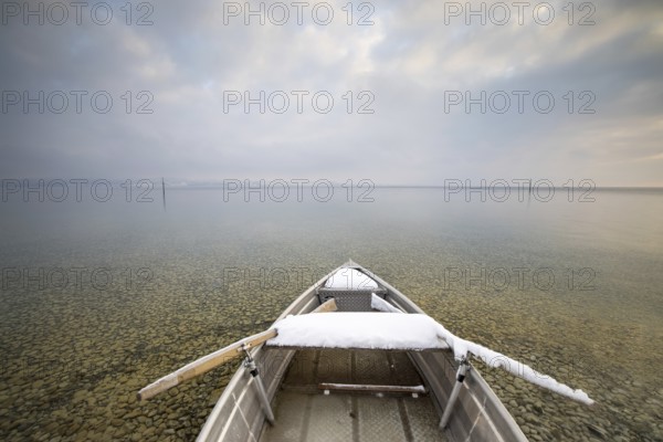 A snow-covered boat on a calm lake with pebbles on the shore under a cloudy sky, Strandbad Hörnle, Constance, Lake Constance, Baden-Württemberg, Germany, Europe