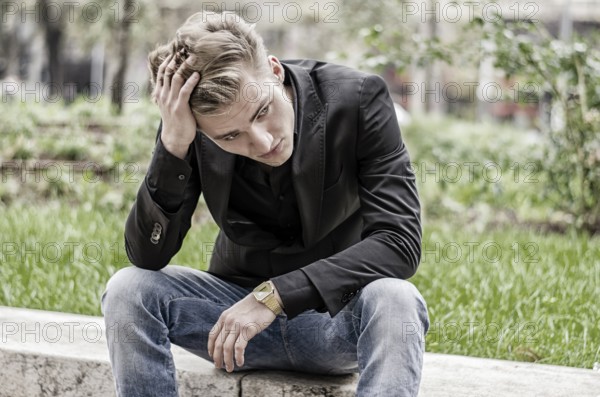 Close up Disappointed Young White Man Sitting at Street Side with Grassy Landscape at the Background