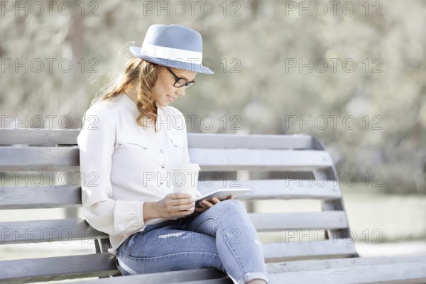 Happy young student with a tablet and a disposable coffee cup sitting on a bench and reading in a summer park