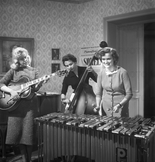Trio 1956. The photograph, taken in 1956, captures three young women in a band, each immersed in their performance. One strums an electric guitar, her fingers poised over the strings with confidence, while another stands beside a double bass, plucking its deep notes with focus. The third plays a xylophone, her mallets striking the wooden bars in a lively rhythm. The scene blends the bold, emerging sound of mid-20th-century music with an unconventional lineup, showcasing a striking mix of instruments and the women's evident talent against a backdrop of 1950s style. Kristoffersson ref BS4-3