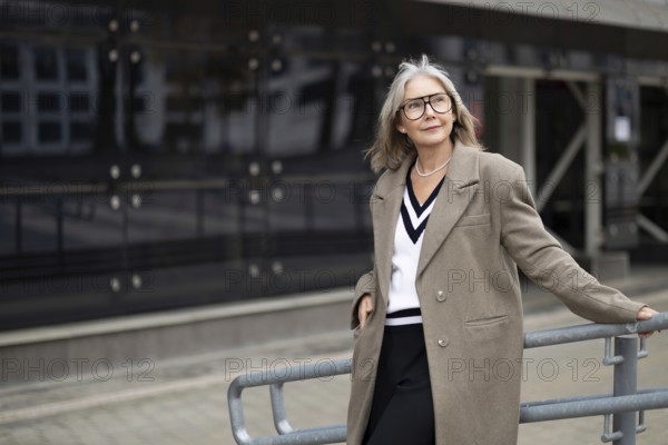 An older woman with gray hair and glasses exudes confidence while leaning against a railing in front of a sleek modern building. She wears a chic outfit that showcases her elegant style