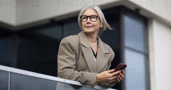 The stylish elderly woman stands elegantly on a balcony, holding her phone and gazing thoughtfully into the distance, bathed in warm afternoon light