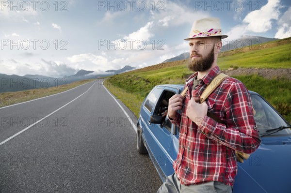 Attractive bearded man in a plaid red shirt and hat with a backpack. Stands next to a retro car on the background of a picturesque road in the mountains at sunset. Easy travel, hitchhiking