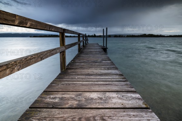 A long wooden footbridge leads into the water of a lake under a dark, cloudy sky, Naturfreundehaus, Markelfingen, Untersee, Lake Constance, Kreis Konstanz, Baden-Württemberg, Germany, Europe