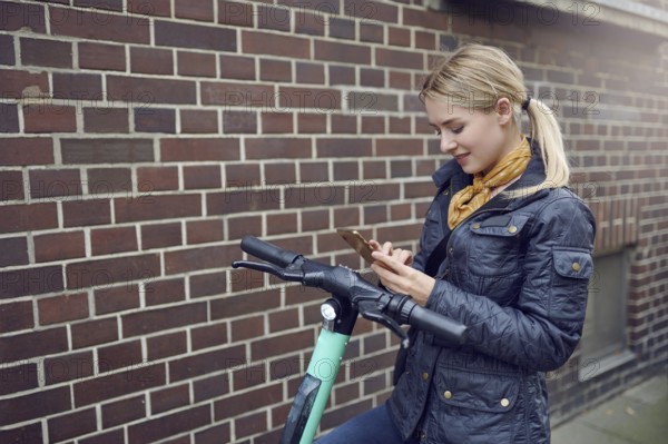 Trendy young blond woman in leather jacket standing at her e-scooter booking a ride, ecological transportation concept with copy space