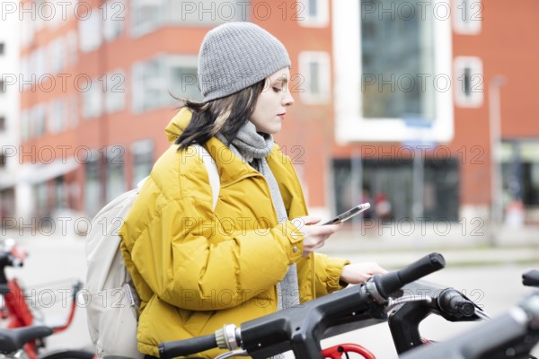 Woman in yellow jacket scrolling on her smartphone next to parked rental bike in the city, environmentally friendly