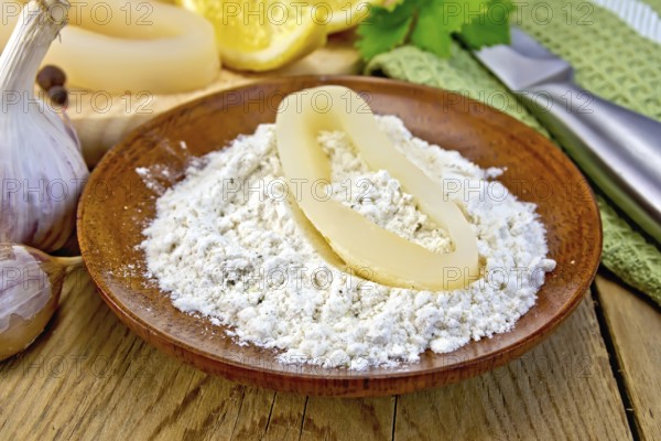 Raw squid rings in flour on a plate, garlic, knife, lemon, napkin on the background of wooden boards on a blackboard