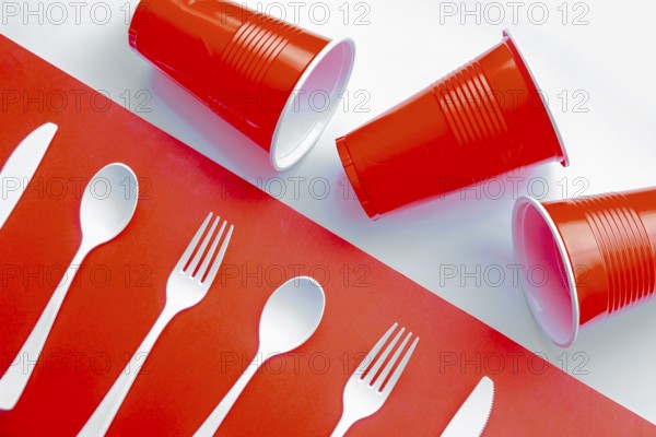 Horizontal view of single use white plastic cutlery and red plastic glass on a red and white background. Concept: Ban single use plastic