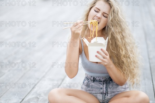 Happy young woman with long curly hair holding a lunch box and eating noodles from a Chinese snack bar with chopsticks