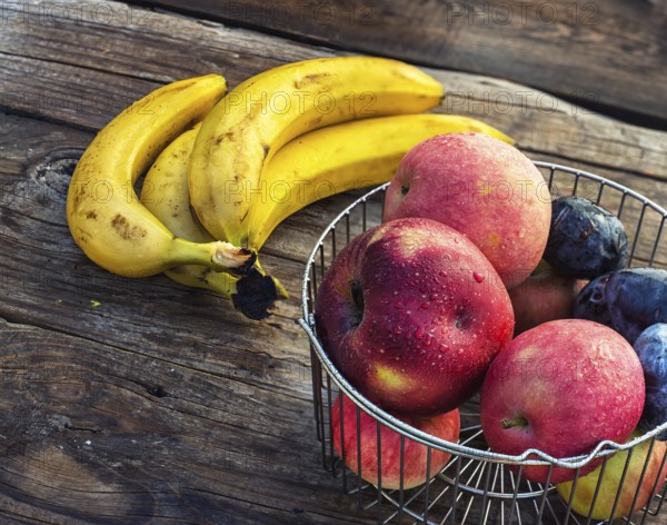 Ripe apples, bananas and plums in metal basket on wooden background