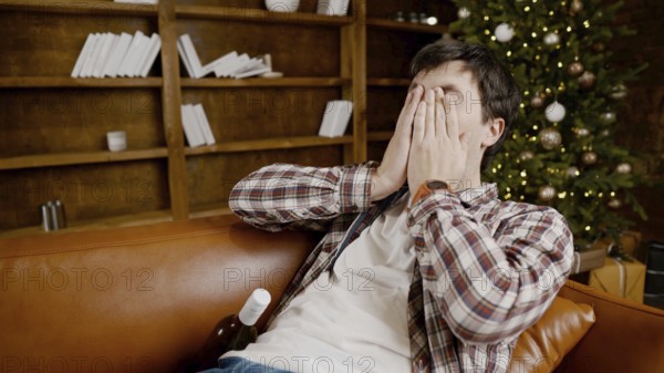 Young male alcoholic requires medical and psychological help. Man addicted alcohol and cannot stay on couch at home on Christmas and New Year Eve. Drunk guy drink wine from bottle near Christmas tree