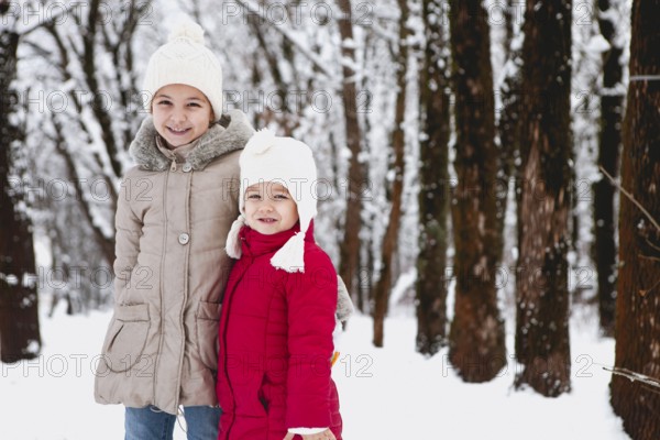 Cheerful little girls in warm clothes smiling and looking at camera while standing on snowy path in winter forest