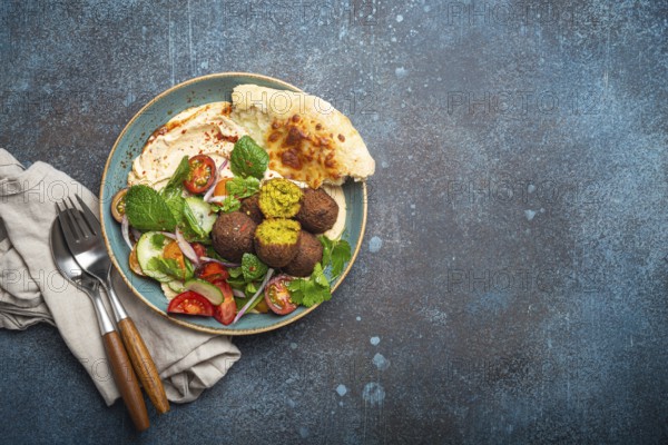 Middle Eastern Arab meal with fried falafel, hummus, vegetables salad with fresh green cilantro and mint leaves, pita bread in ceramic bowl on stone rustic background top view, space for text, food photography