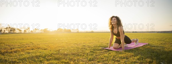 Person smiles while in a yoga pose on a mat, lit by the morning sun, in a field