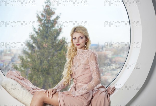 Portrait of young beautiful caucasian blond girl in gorgeous dress near the window