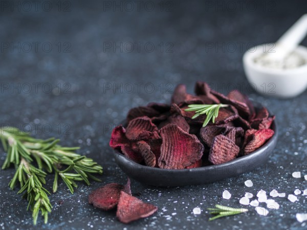 Baked beet slices with coarse sea salt scented rosemary. Vegan diet food idea and recipe. Healthy homemade beetroot chips in plate and ingredients on dark blue background. Copy space