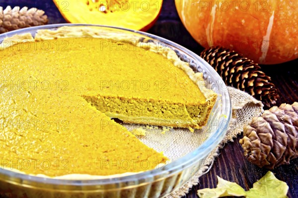 Pumpkin pie in a glass pan on sackcloth, pumpkins, pine cones and maple leaf on the background of dark wood planks