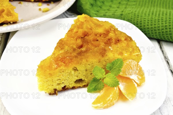 Piece of tart with mandarin, mint, mandarin slices in a white plate, towel on a background of wooden boards
