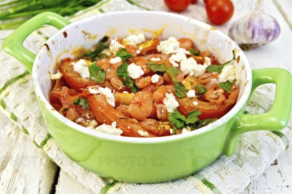 Shrimp and tomatoes baked with feta cheese in a roasting pan on a towel, parsley and garlic on a wooden boards background