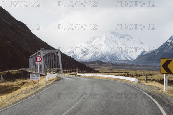 Road leading to a bridge with snow-capped mountains and cloudy sky in the background, Hooker Valley Track, New Zealand, Oceania