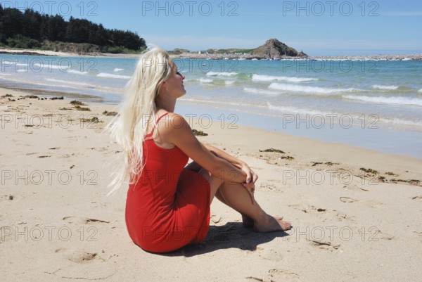 Woman at beach, A woman in a red dress sits on the sandy beach in Erquy, facing the sea under a clear blue sky