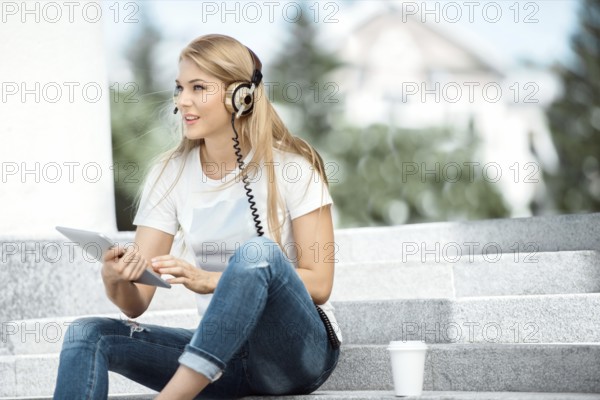 Happy young woman with vintage headphones and a cup of coffee to go, surfing the internet on a tablet PC, listening to music and sitting on a staircase in front of an urban background