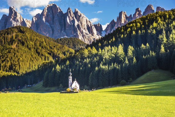 Dolomites, Tirol. Sunny autumn day. The famous symbol of the valley Val di Funes - church of Santa Maddalena. Rocky peaks and forested mountains surrounded by green Alpine meadows