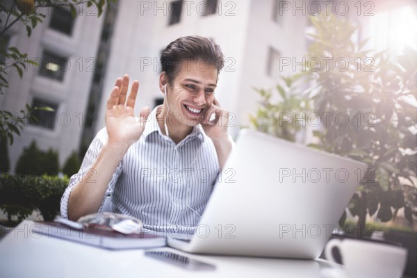 Happy handsome businessman looking at computer screen while typing something on laptop keyboard during work break outdoors, businessman with headphones looking at the notebook sitting in cafe working