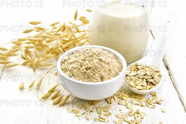 Flour oat in bowl, milk in a jug, oatmeal in spoon, oaten stalks on the background of light wooden board